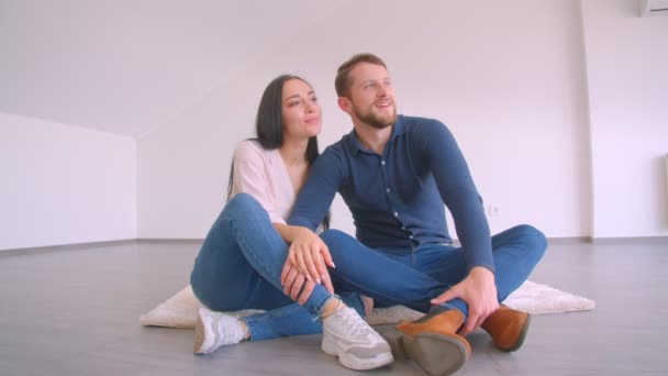Progressives kaukasisches Paar, das auf dem Boden der neuen leeren Wohnung sitzt, sich umarmt und küsst, glücklich auf weißem Wandhintergrund — Stockvideo
