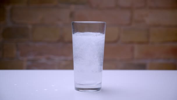 Tiro close-up de vidro transparente em pé sobre a mesa branca com água e pílula sendo jogado nele — Vídeo de Stock