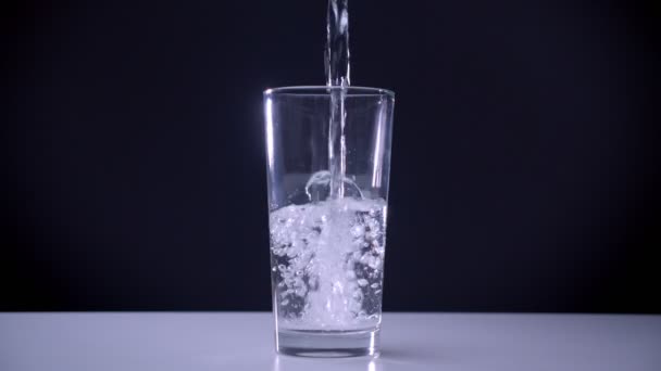 Съемка крупным планом прозрачного стекла, стоящего на белом столе, и газированной воды, льющейся в него на изолированном фоне — стоковое видео