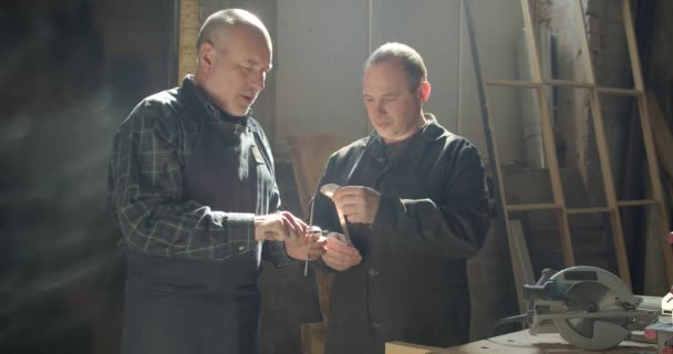 Dos maestros de la carpintería de pie en la fabricación de madera discutiendo el equipo de observación en tachuelas . — Vídeo de stock