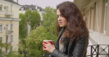 Muhteşem yeşil şehir manzarası zevk ve sıcak çay içme balkonda duran deri ceketgenç esmer kız.