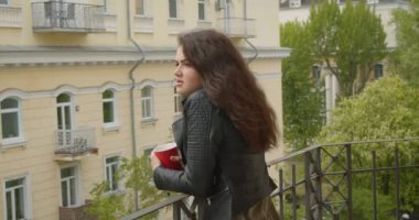 Deri ceketgüzel esmer kız balkondan yeşil şehir görünümünde izlerken ve rüya gibi ve sakin olmak sıcak çay içme.