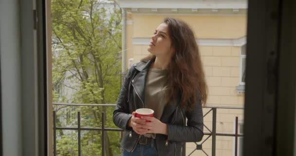 Jolie fille brune en veste en cuir debout sur le balcon avec vue sur la ville verte et boire du thé chaud étant rêveuse et calme . — Video