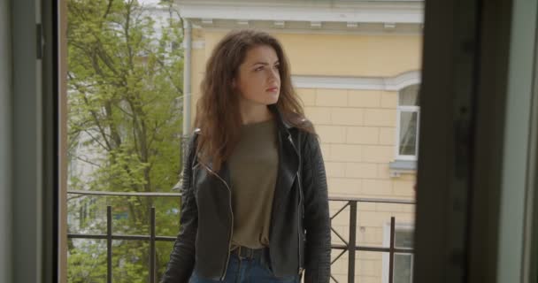 Pretty brunette girl in leather jacket standing on balcony with green city view smiling into camera being glad and happy. — Stock Video