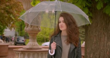 Yeşil şehir parkında kameraya güzel ve sevinçle gülümseyen şemsiyeli beyaz esmer kadın portresi.