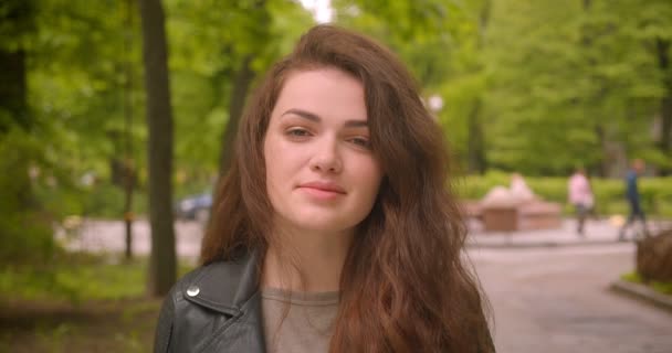 Close-up shot van Kaukasische brunette vrouwelijke horloges in de camera met rustige glimlach in het groene stadspark. — Stockvideo