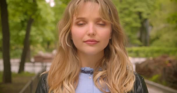 Jolie étudiante blonde caucasienne regardant modestement et calmement dans la caméra dans le parc de la ville verte . — Video