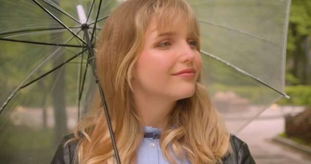 Jolie étudiante blonde caucasienne avec parapluie souriant timidement dans la caméra dans le parc de la ville verte . — Video