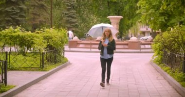 Güzel beyaz sarışın öğrenci şemsiye ile yeşil park boyunca doğanın güzelliğini gözlemleyerek yürüyüş kameraya gülümsüyor.
