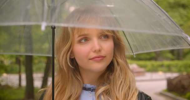 Pretty kaukasiska blonda student Smiles glatt i kameran stående under paraply i den gröna stadsparken. — Stockvideo