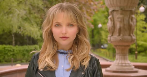 Bella studentessa bionda caucasica sorridente in macchina fotografica vicino alla fontana nel parco verde della città . — Video Stock