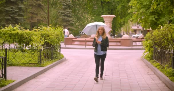 Mooie blanke blonde student met paraplu wandelen langs Green Park het observeren van de schoonheid van de natuur glimlacht in de camera. — Stockvideo