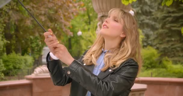 Pretty caucasian blonde student opens the umbrella preparing for rain being glad and optimistic in the green city park. — Stock Video