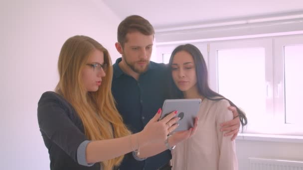 Риэлтор показывает молодой кавказской паре квартиру на продажу, демонстрируя и обсуждая возможности с помощью планшета . — стоковое видео