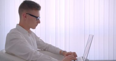 Beyaz bir odada kapalı koltukta oturan dizüstü bilgisayar kullanarak gözlük genç yakışıklı kafkas işadamıcloseup yan görünüm portresi