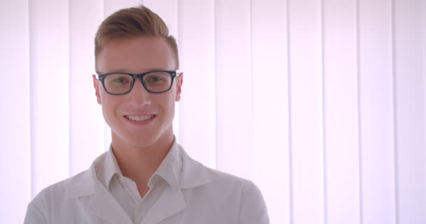 Närbild porträtt av unga stilig kaukasiska affärsman i glasögon tittar på kamera leende lyckligt stående inomhus i ett vitt rum — Stockvideo