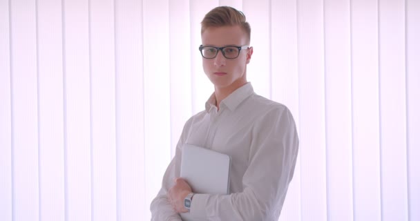 Retrato de primer plano de un joven guapo hombre de negocios caucásico con gafas sosteniendo un portátil en el interior de una habitación blanca — Vídeo de stock
