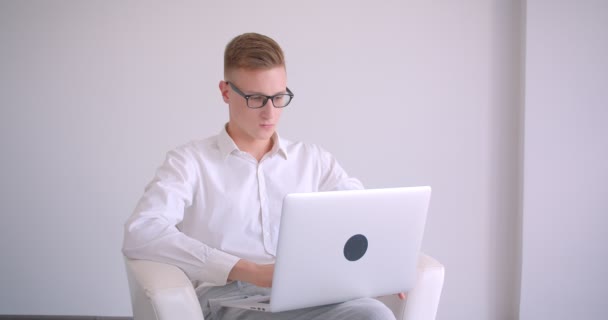 Retrato de primer plano del joven hombre de negocios caucásico guapo en gafas usando el portátil mirando a la cámara sonriendo felizmente sentado en el sillón dentro de una habitación blanca — Vídeos de Stock