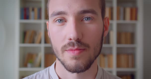 Primer plano retrato de joven guapo caucásico estudiante masculino mirando a la cámara sonriendo alegremente en la biblioteca de la universidad con estanterías en el fondo — Vídeos de Stock