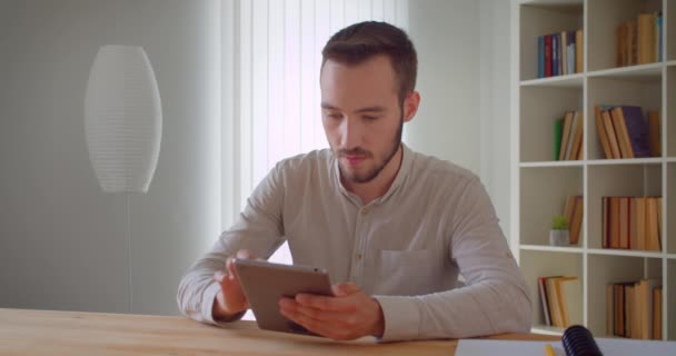 Nahaufnahme Porträt eines jungen gutaussehenden kaukasischen Geschäftsmannes, der das Tablet in der Wohnung mit Bücherregalen im Hintergrund benutzt — Stockvideo