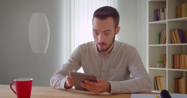 Nahaufnahme Porträt eines jungen gutaussehenden kaukasischen Geschäftsmannes, der in der Wohnung auf dem Tablet surft — Stockvideo
