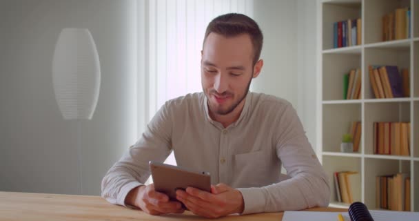 Primer plano retrato de joven hombre de negocios caucásico guapo usando la tableta mirando a la cámara sonriendo alegremente en el interior del apartamento — Vídeos de Stock