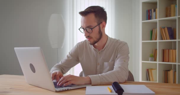 Genç yakışıklı beyaz işadamının dizüstü bilgisayarda yazarak dairenin iç mekanındaki kameraya bakan yakın çekim portresi — Stok video