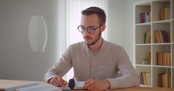 Retrato de primer plano de un joven guapo estudiante caucásico en gafas que estudia y usa la tableta tomando notas mirando la cámara en la biblioteca de la universidad — Vídeo de stock