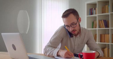 Genç yakışıklı beyaz işadamının dairenin içinde dizüstü bilgisayarı kullanarak telefon görüşmesi yaparken ki yakın çekim portresi