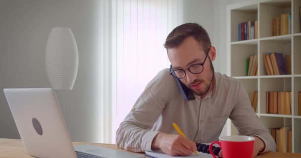 Close-up portret van jonge knappe Kaukasische zakenman met een telefoontje met behulp van de laptop binnenshuis in het appartement — Stockvideo