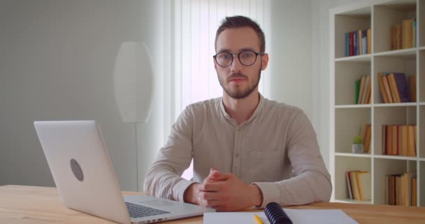 Close-up portret van jonge knappe Kaukasische zakenman kijken naar camera zittend in de voorkant van de laptop binnenshuis in het appartement — Stockvideo