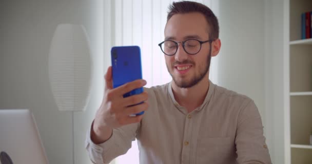 Closeup portré fiatal csinos kaukázusi businessmantaking selfies telefonon mosolyogva boldogan ül a laptop előtt bent a lakásban beltéri — Stock videók