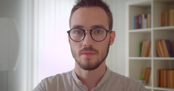 Gözlüklü genç yakışıklı beyaz erkek öğrencinin kapalı dairedeki kameraya bakan yakın çekim portresi — Stok video
