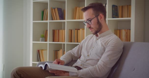 Genç yakışıklı beyaz işadamının dairenin içinde kanepede oturan kameraya bakan bir kitap okuma closeup portre — Stok video