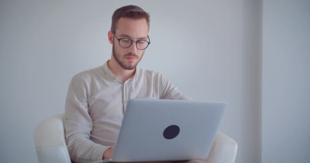 Κοντινό πλάνο του νεαρού όμορφος Καυκάσιος επιχειρηματίας που δακτυλογραφεί στο φορητό υπολογιστή κοιτάζοντας την κάμερα χαμογελώντας ευτυχισμένα καθισμένος στην πολυθρόνα στο λευκό διαμέρισμα — Αρχείο Βίντεο