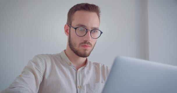 Zbliżenie portret młodego przystojnego kaukaski biznesmen wpisując na laptopie patrząc na aparat w pomieszczeniu w białym mieszkaniu — Wideo stockowe