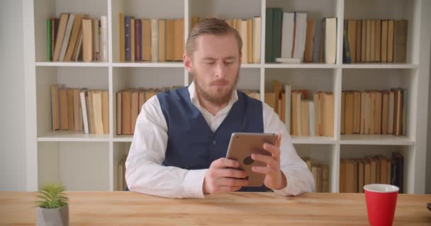 Крупним планом портрет молодого кавказького бізнесмена, який використовує планшет, дивлячись на камеру в офісі в приміщенні з книжковими полицями на фоні — стокове відео