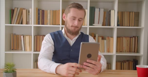 Retrato de close-up de jovem empresário caucasiano que tem uma chamada de vídeo no tablet no escritório dentro de casa com estantes no fundo — Vídeo de Stock