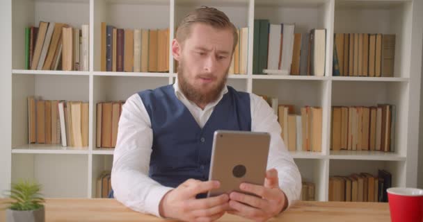 Close-up portret van jonge Kaukasische zakenman met een formeel videogesprek op de Tablet in het kantoor binnenshuis met boekenkasten op de achtergrond — Stockvideo