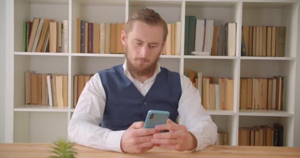 Крупный план портрета молодого кавказского бизнесмена, пользующегося телефоном и весело улыбающегося в офисе в помещении с книжными полками на заднем плане — стоковое видео