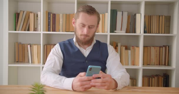 Retrato de primer plano del joven hombre de negocios caucásico usando el teléfono y sonriendo alegremente en la oficina en el interior con estanterías en el fondo — Vídeos de Stock