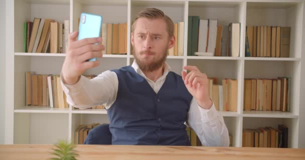 Närbild porträtt av unga kaukasiska businessmantaking selfies på telefonen på arbetsplatsen inomhus med bokhyllor på bakgrunden — Stockvideo