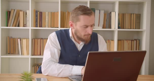 Крупним планом портрет молодого Кавказького бізнесмена з використанням ноутбука і телефонний дзвінок в офісі в приміщенні з книжкові полиці на задньому плані — стокове відео