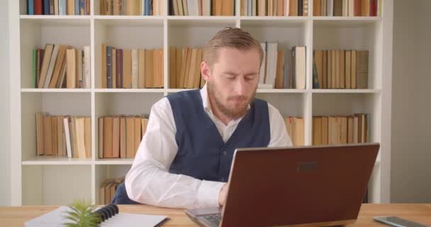 Портрет крупным планом молодого кавказского бизнесмена, использующего ноутбук и делающего заметки на рабочем месте в помещении с книжными полками на заднем плане — стоковое видео