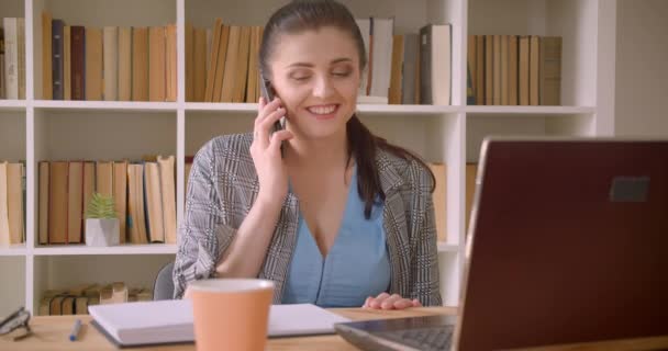 Närbild shoot av unga kaukasiska affärskvinna med hjälp av den bärbara datorn och pratar känslomässigt på telefon l i bibliotekets kontor inomhus — Stockvideo