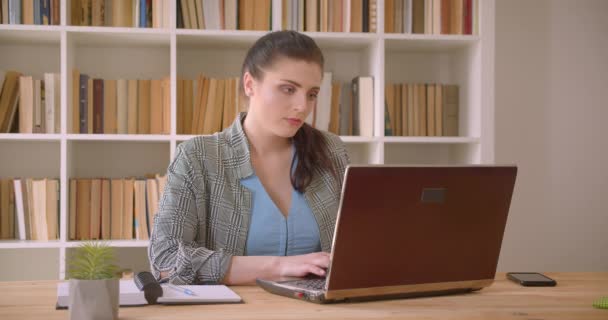 Nahaufnahme einer jungen kaukasischen Geschäftsfrau, die mit dem Laptop in die Kamera blickt und im Bibliotheksbüro drinnen glücklich lächelt — Stockvideo