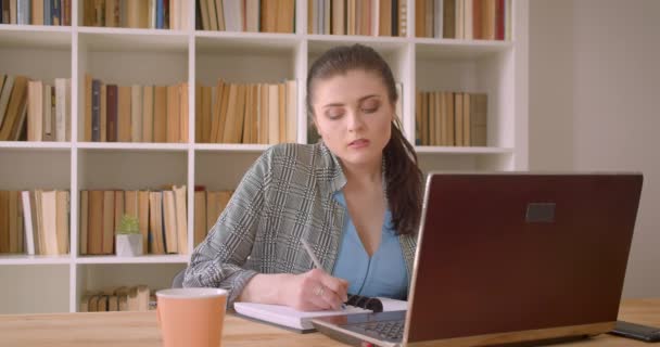 Närbild shoot av unga kaukasiska affärskvinna med hjälp av den bärbara datorn gertting varm och trött i bibliotekets kontor inomhus — Stockvideo