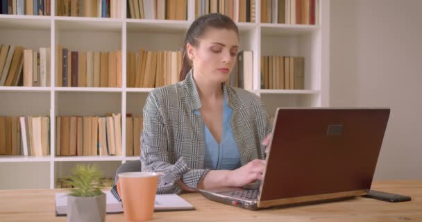 Dizüstü bilgisayar üzerinde çalışan genç beyaz iş kadınının kapalı kütüphane ofisinde güvenle gülümseyen kameraya bakarak yakın çekim çekimi — Stok video