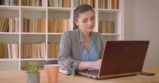 Tiro de close-up de jovem mulher de negócios caucasiana usando o laptop e tomar notas no escritório da biblioteca dentro de casa — Vídeo de Stock