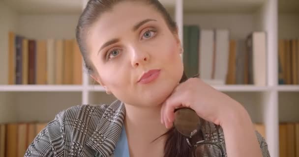 Nahaufnahme einer jungen kaukasischen Geschäftsfrau, die gelangweilt in die Kamera blickt, im Bibliotheksbüro mit Bücherregalen im Hintergrund — Stockvideo